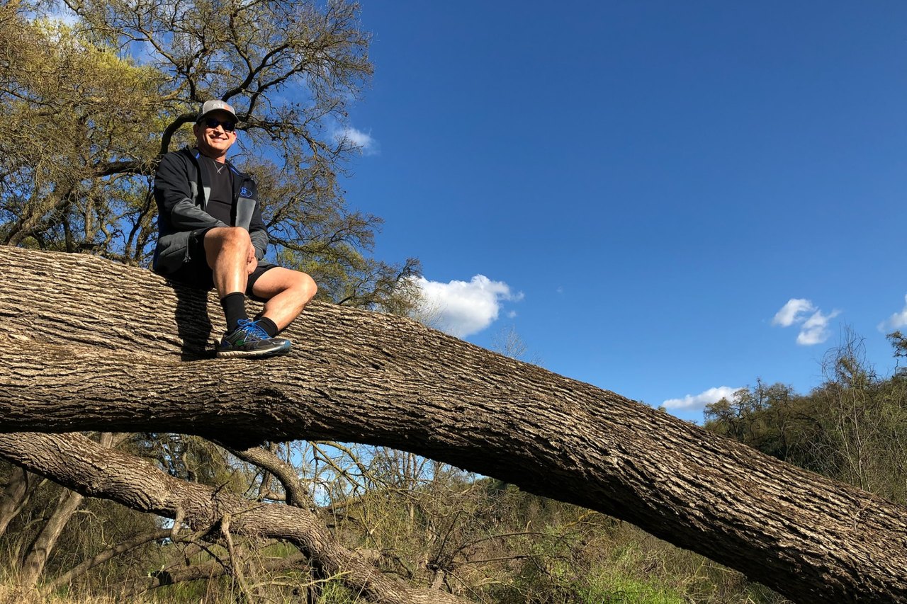 Todd Valdez sitting on the wide trunk of a large fallen oak, outdoors under an open blue sky.