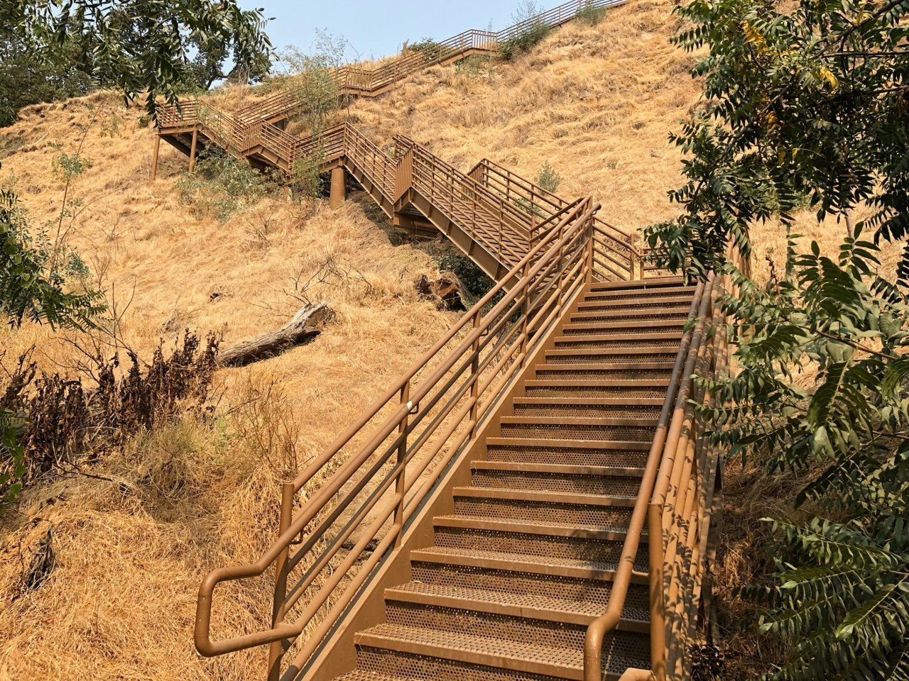 A long metal staircase with railings climbing a dry grass hillside, switching back toward a walkway at the top.