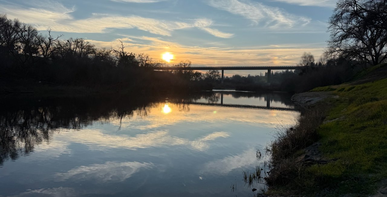 Sunset over a calm river, with a long bridge crossing in the distance and the sky reflected in the water.